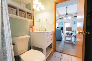 A bathroom with a toilet, sink, and shelves with baskets. There is a door leading to a living area with a ceiling fan, a couch, and a coffee table. - Charming 4-bedroom house.  Walking distance to the Beach and Restaurants! (Kill Devil Hills)
