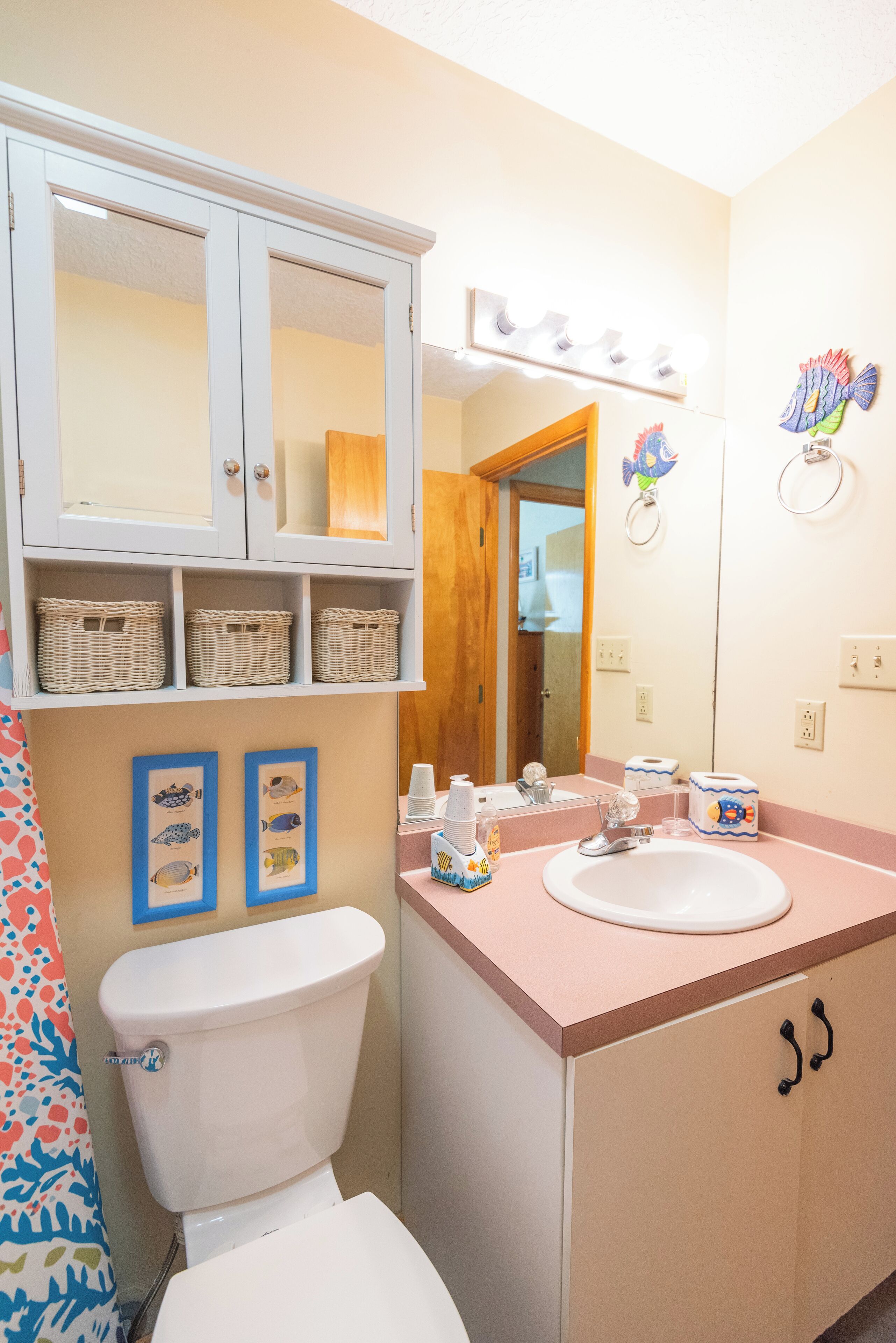 Une salle de bain avec des toilettes blanches, un lavabo surmonté d’un miroir et un meuble avec des paniers en osier.