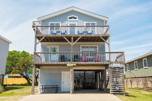 A two-story building with a balcony on the upper floor, a garage door, and a small outdoor seating area. - Charming 4-bedroom house.  Walking distance to the Beach and Restaurants! (Kill Devil Hills)
