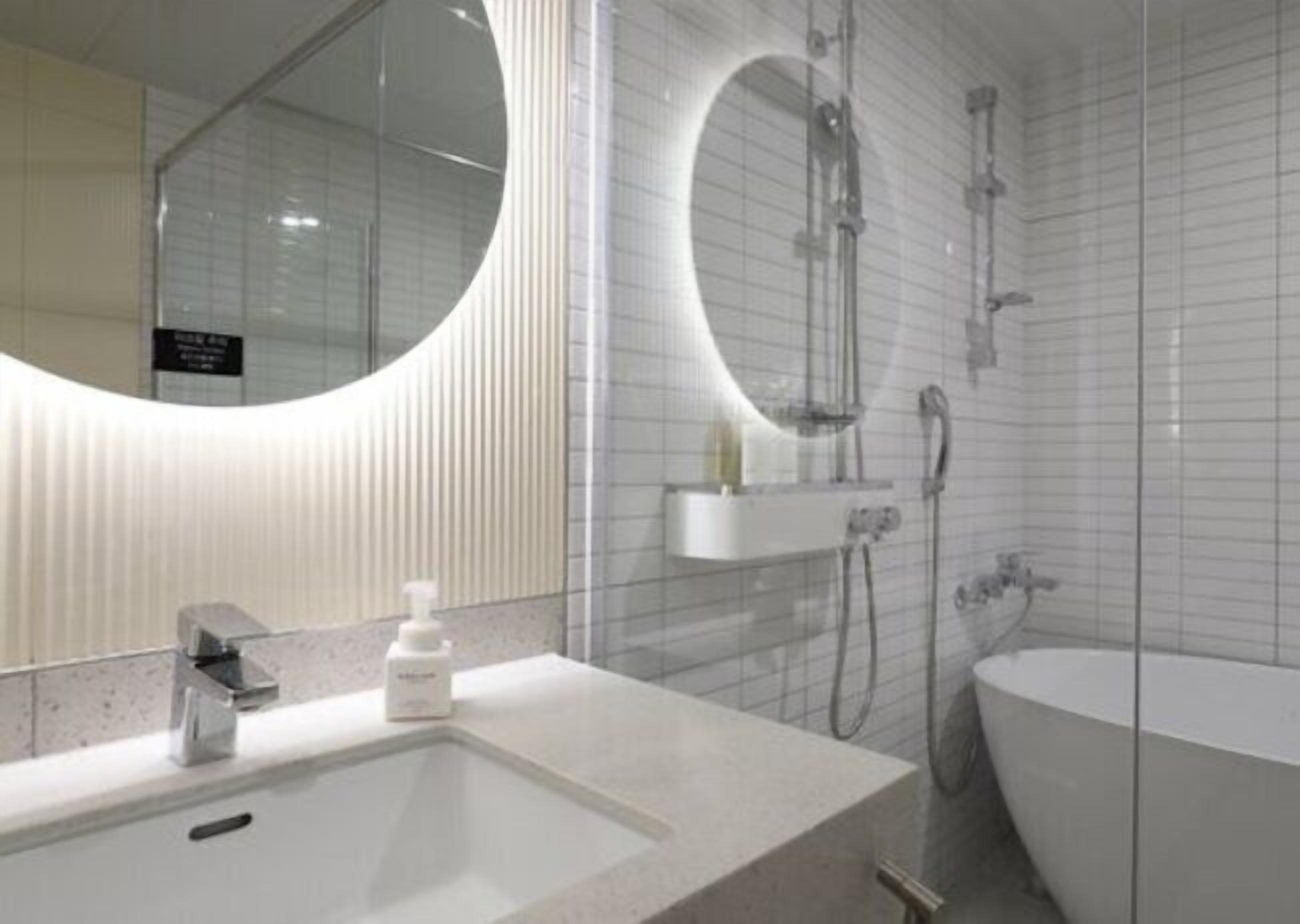 A modern bathroom with a large oval mirror, a rectangular sink, a handheld showerhead, and a standalone bathtub.