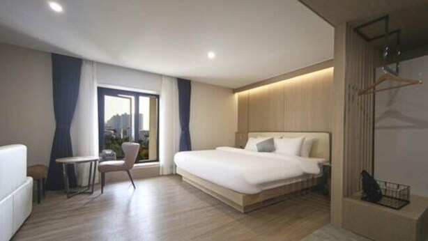 A modern hotel room with a large bed, a small table, a chair, and a view of the city through the window. - H AVENUE (Busan)