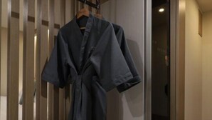A dark-colored robe hanging on a wooden hanger in a hallway with a wooden door and a light switch on the wall. - H AVENUE (Busan)