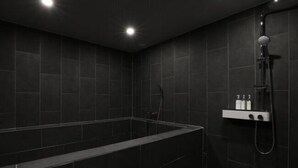 A modern bathroom with a large rectangular bathtub, dark tiled walls, and a shower area with a handheld showerhead. - H AVENUE (Busan)