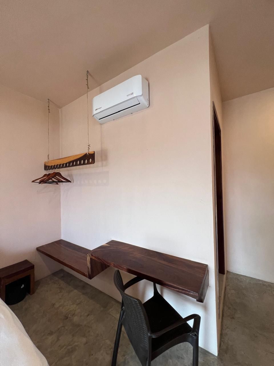 Quarto pequeno e simples com um banco de madeira, uma cadeira preta, um suporte de madeira suspenso e um aparelho de ar condicionado branco na parede.