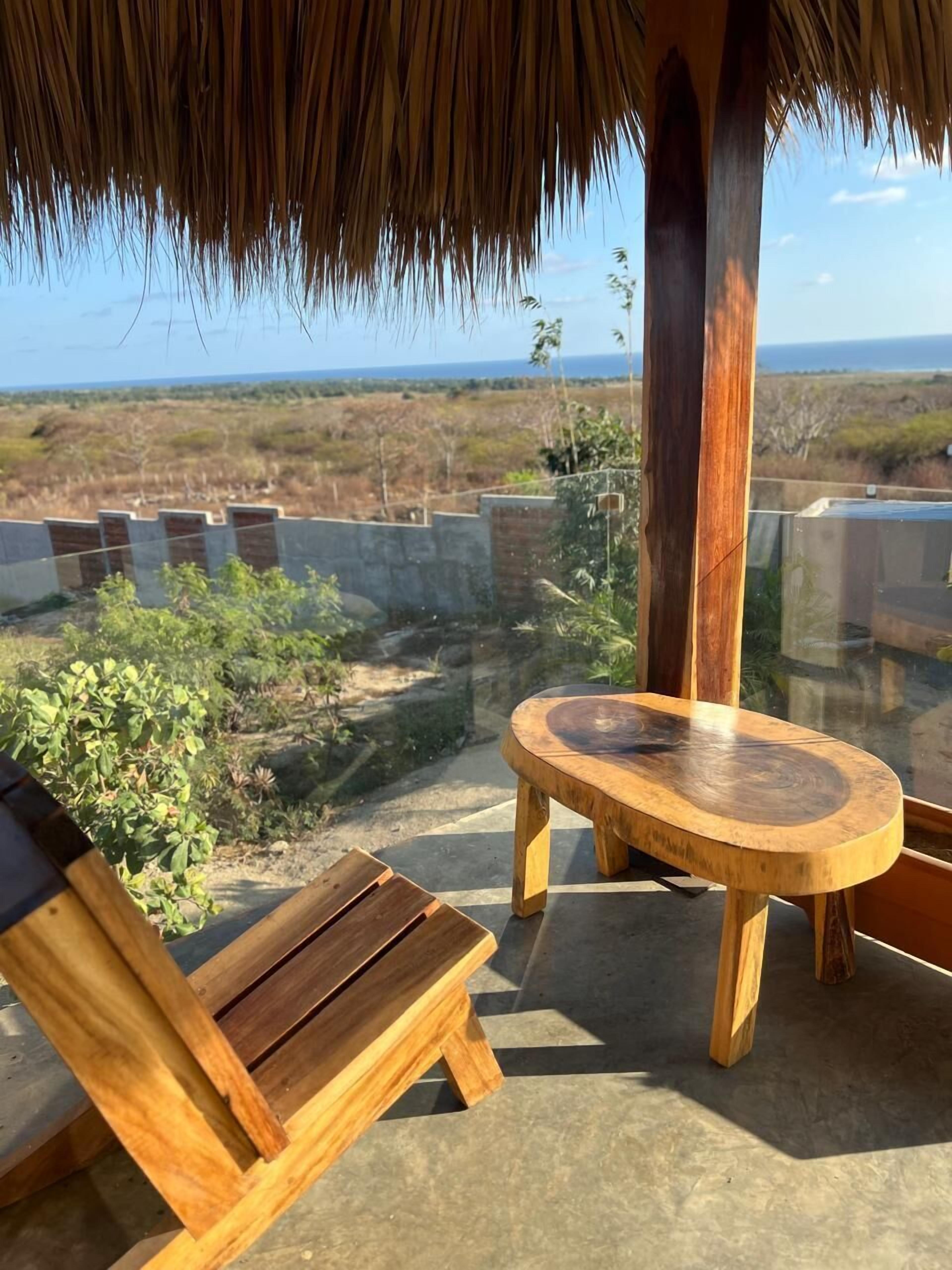 A wooden bench and chair under a thatched roof, overlooking a coastal landscape.