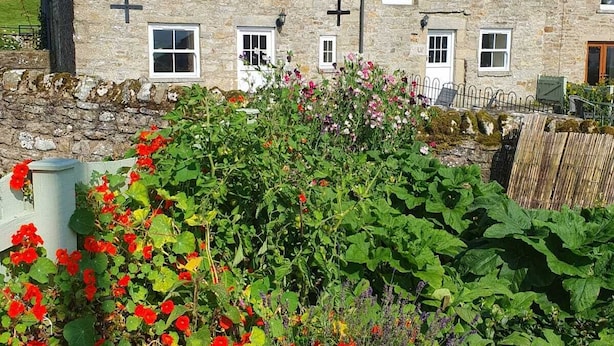 A stone building with multiple windows and a cross on the wall, surrounded by a garden with various plants and flowers. - Lonin Cottage. Unwind in our cozy cottage, surrounded by scenic walks. (Barnard Castle)
