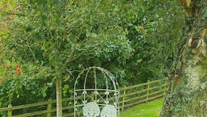 A garden with a white metal bench under a tree, surrounded by greenery and a wooden fence. - Lonin Cottage. Unwind in our cozy cottage, surrounded by scenic walks. (Barnard Castle)