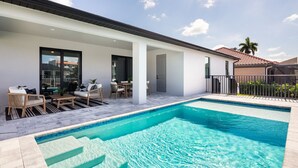 A modern outdoor pool area with a clear blue pool, a covered seating area with chairs and tables, and a view of a neighboring building with a red-tiled roof.