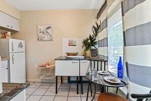 Cocina compacta con refrigerador, una pequeña mesa de comedor y una silla. Se observa una ventana con vista a la calle.