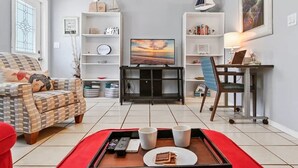 Ein Wohnzimmer mit einer Couch, einem Fernseher, einem Schreibtisch mit Stuhl und einem Bücherregal.