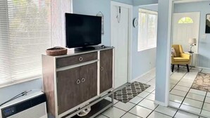 A living room with a TV on a cabinet, a heater, a window with blinds, a door, and a yellow chair. - BEACH OASIS - Walk 2 Beaches, Restaurants & Shops... Pets ok (Deerfield Beach)