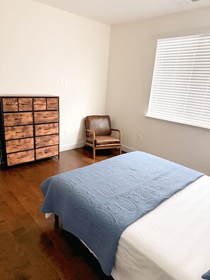 Un dormitorio con una cama, un aparador de madera, una silla y un ventanal con persianas.