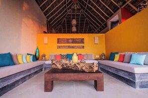Ein Zimmer mit hohen Decken und einem Strohdach, ausgestattet mit zwei Sofas mit bunten Kissen, einem Holztisch und einem Wandbild.