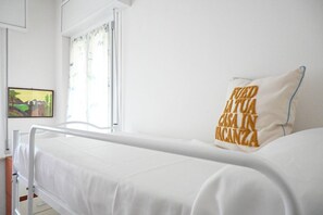 Une chambre avec un lit simple, un lit blanc, une fenêtre avec des rideaux et un oreiller orné d’un motif.