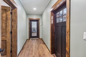 Un corridoio con pavimento in legno, una porta scura con pannelli di vetro e vista su un'area salotto esterna.