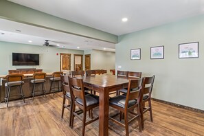 A dining area with a wooden table and chairs, a bar with stools, and a television on the wall.
