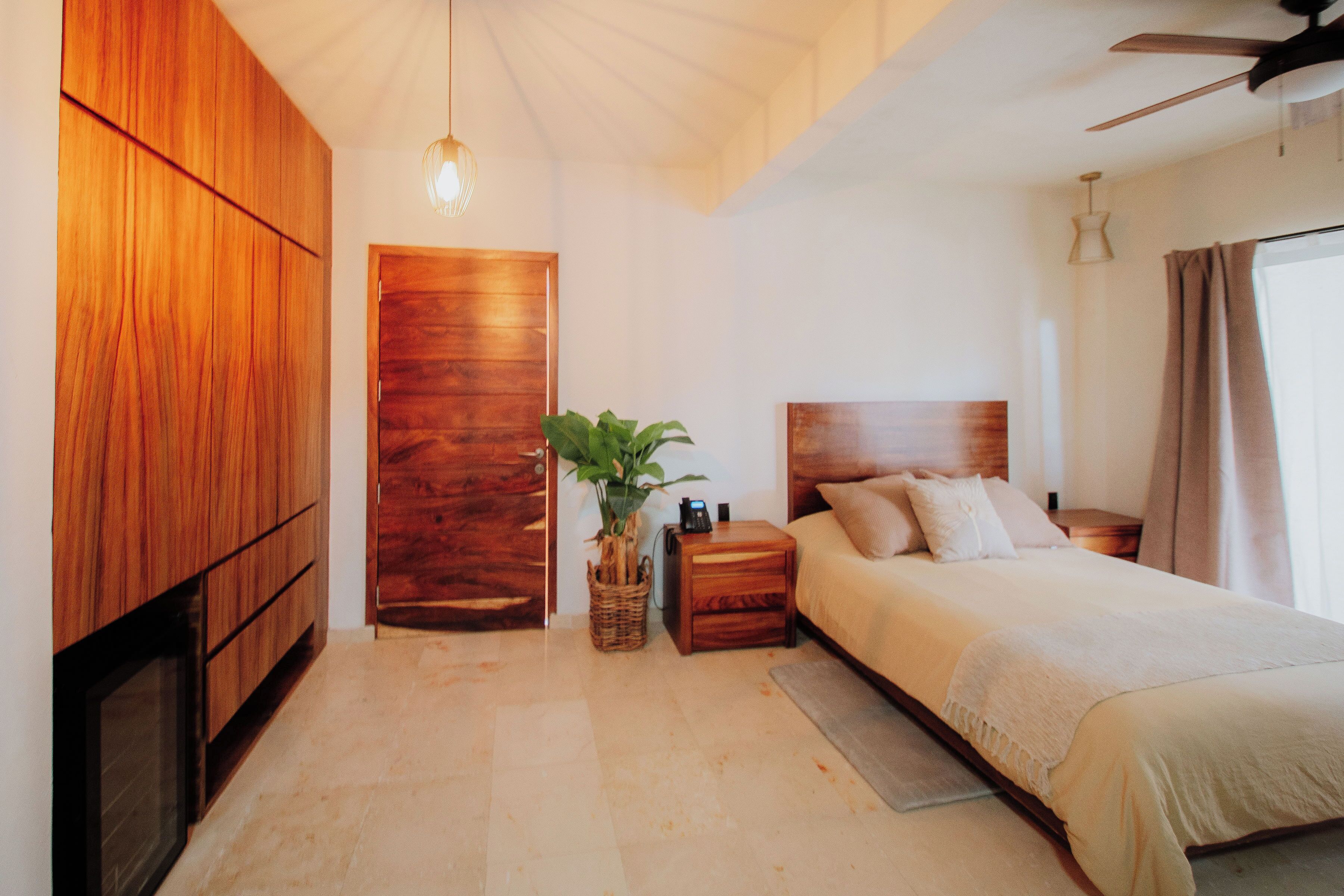 A bedroom with a wooden wardrobe, a bed with a headboard, a nightstand, a lamp, and a ceiling fan.