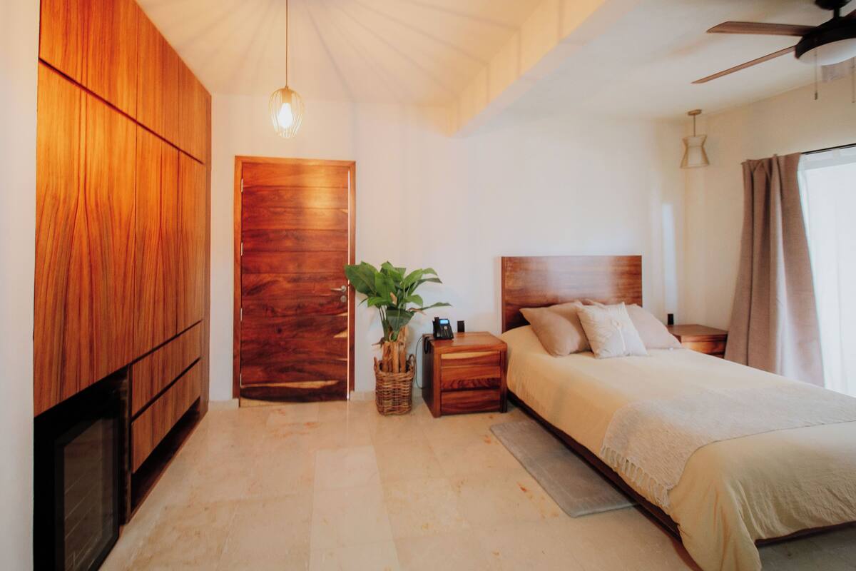 Une chambre avec une armoire en bois, un lit avec une tête de lit, une table de chevet, une lampe et un ventilateur de plafond.