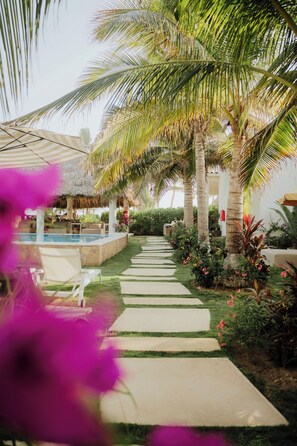Zona de piscina con sillones de descanso, una estructura de techo de paja y palmeras.