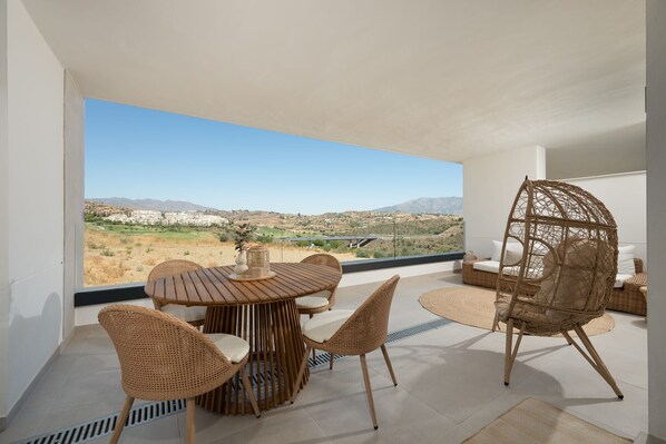 A balcony with a round wooden table, wicker chairs, and a hanging chair. - Casa MLML | Apartment with Mountain View in Mijas (La Cala de Mijas)