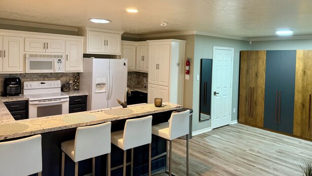 A modern kitchen with white cabinets, a stainless steel refrigerator, a microwave, and a stove. There are two white bar stools and a wooden door in the background.