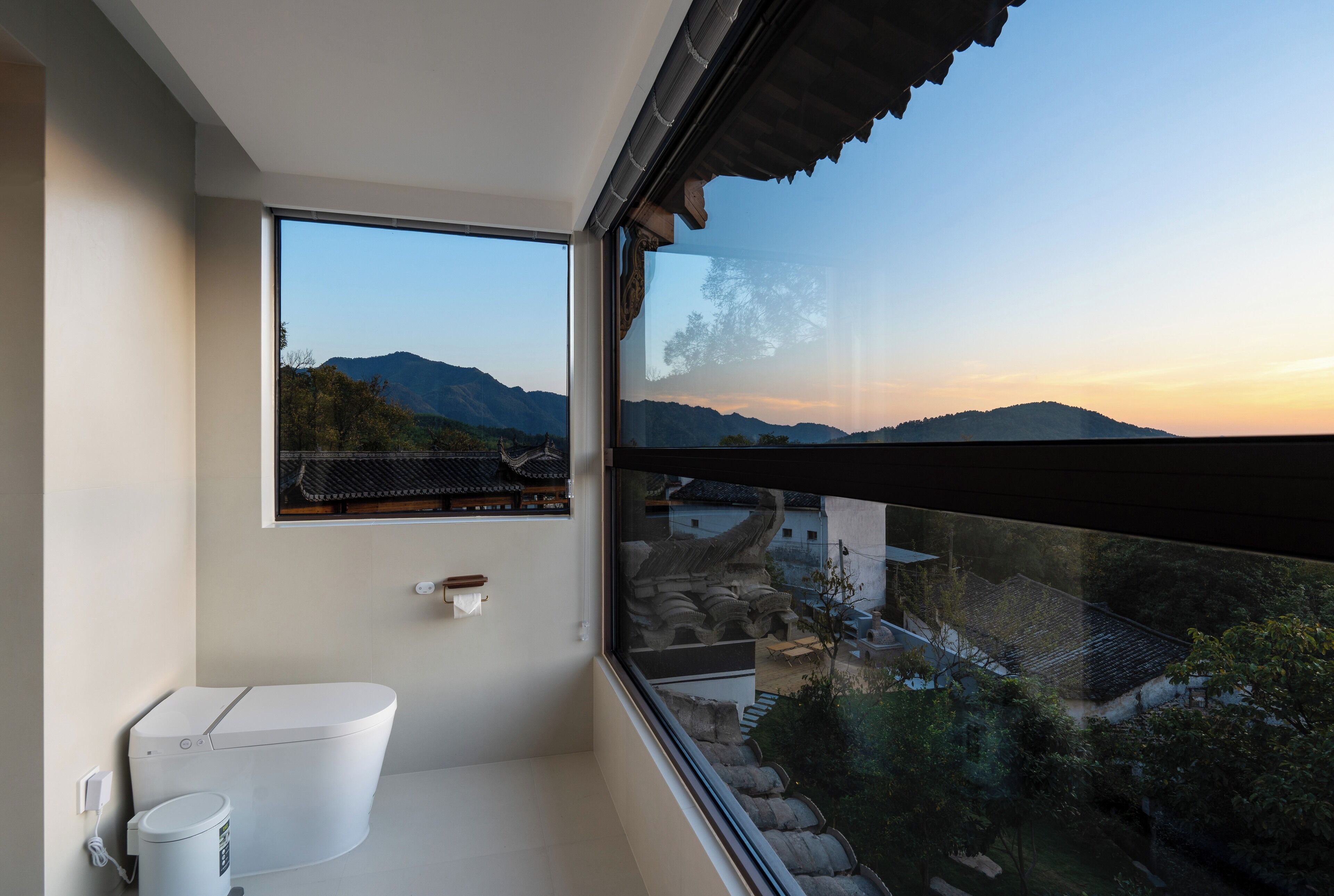 Un baño moderno con inodoro, bidé y un amplio ventanal con vistas a montañas y edificios tradicionales.