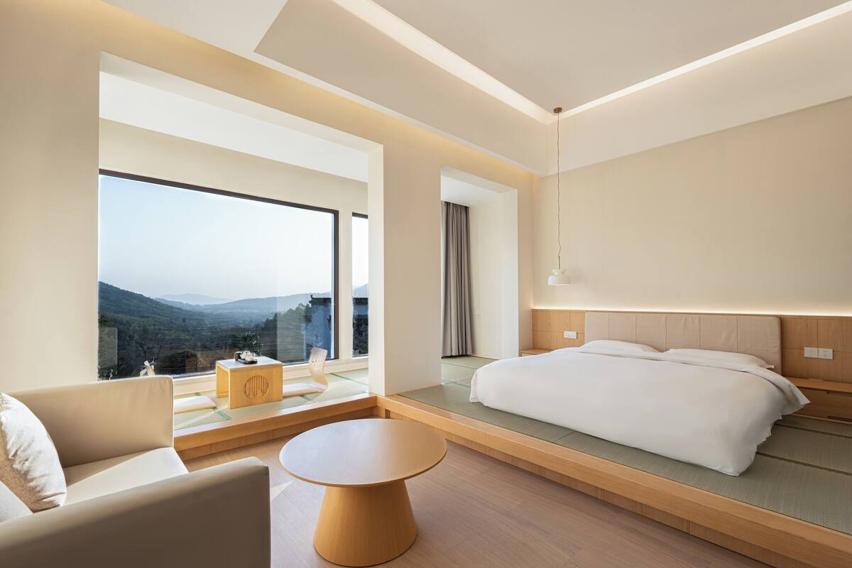 A modern hotel room with a large window offering a mountain view, a bed with a wooden headboard, a small round table, and a sofa.
