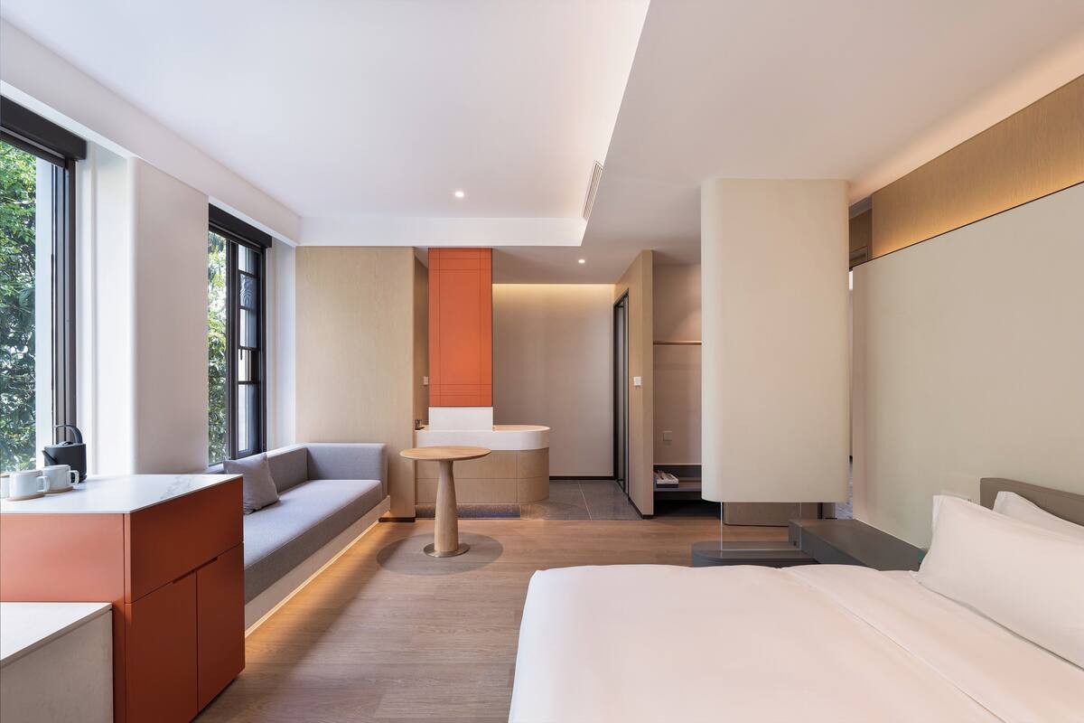 A modern hotel room with a bed, a seating area, a small table, and a view of the outdoors.