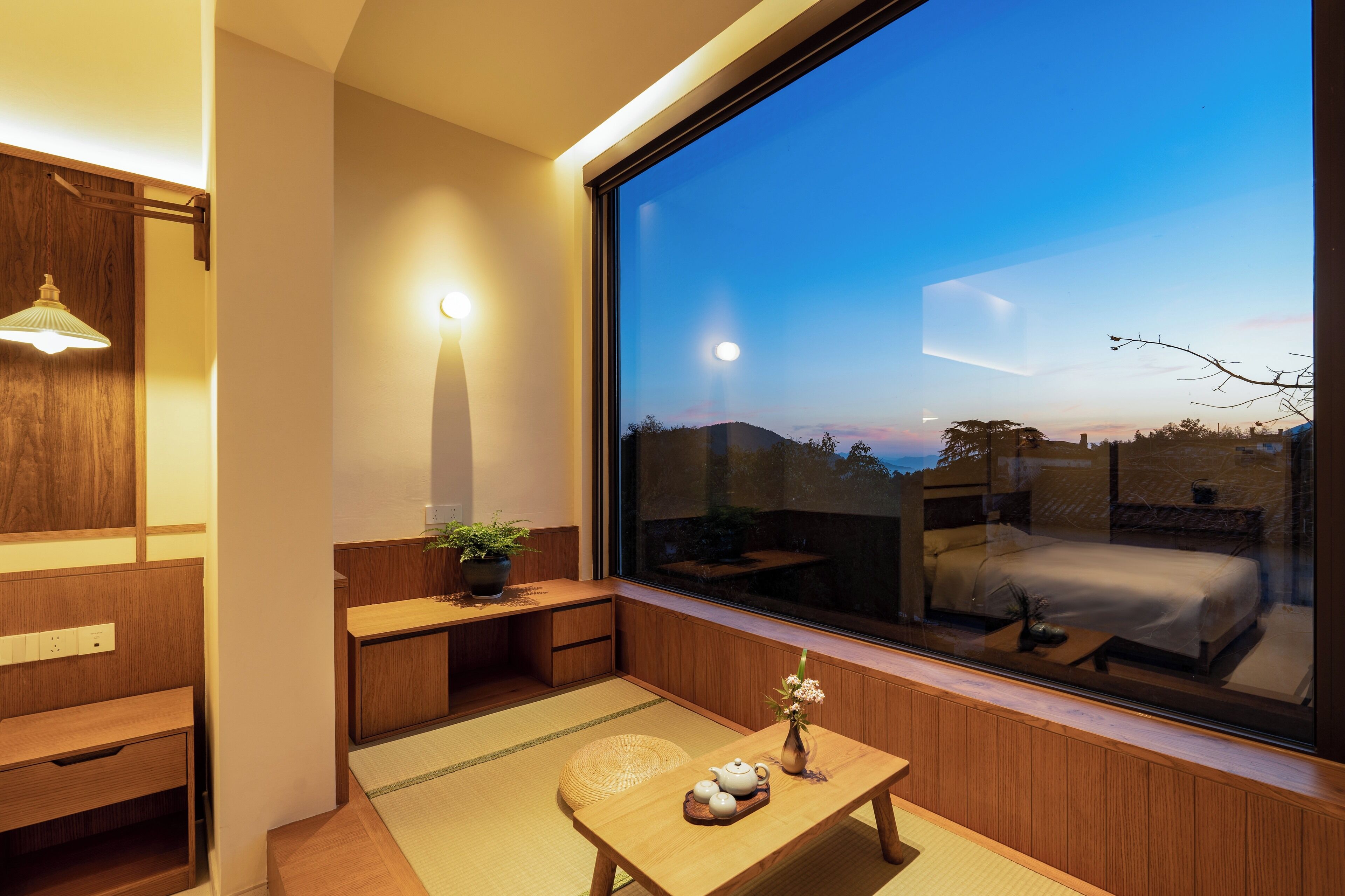 A modern room with a large window, wooden furniture, a small table with a vase and teapot, and a view of a bed and outdoor area.