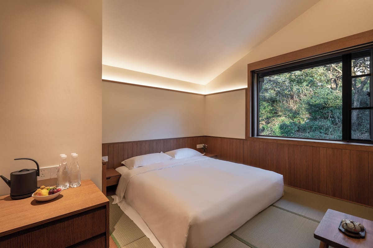 A neatly arranged hotel room with a bed, bedside table, and a window offering a view of greenery.