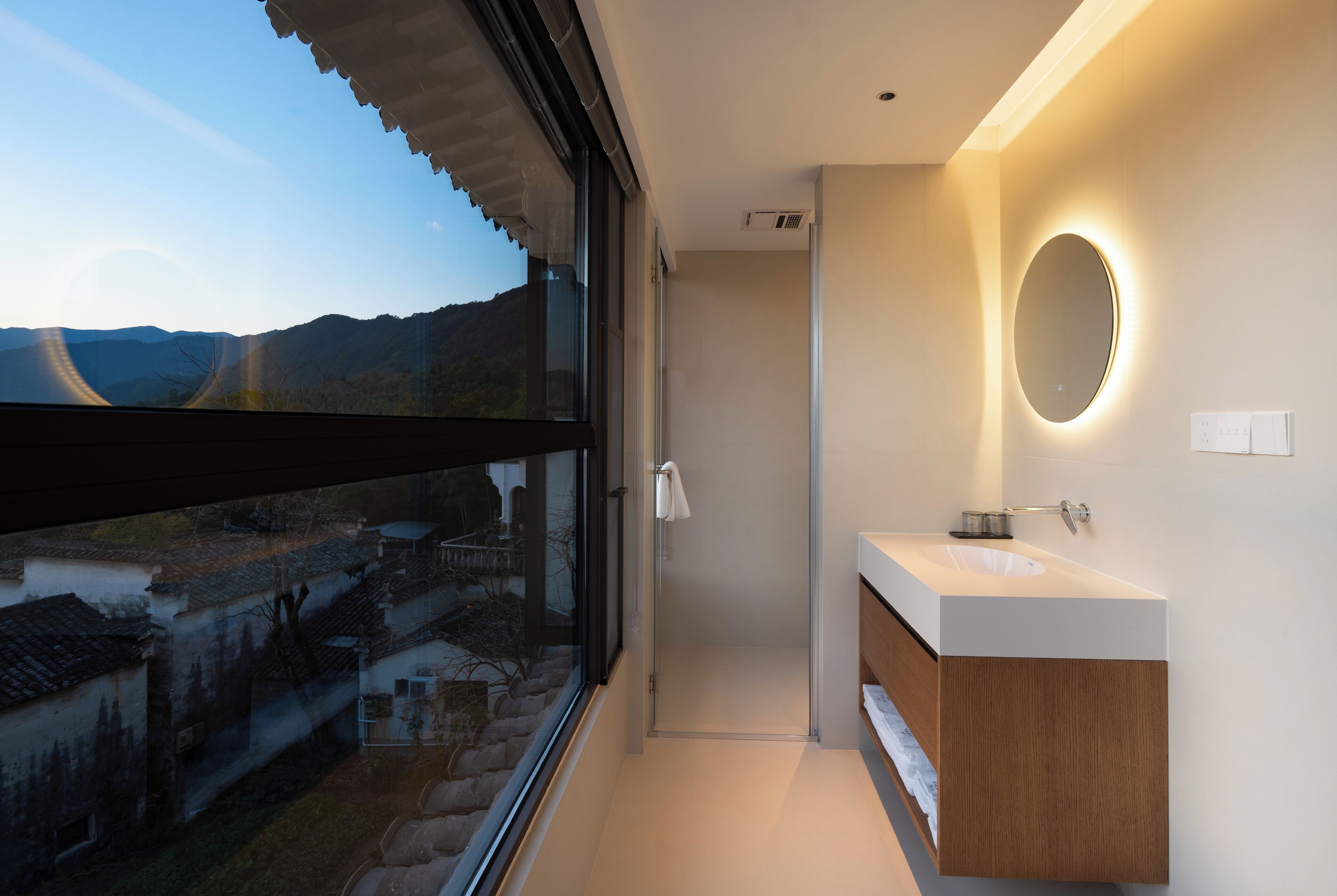 A modern bathroom with a large window, a round mirror, and a white sink.