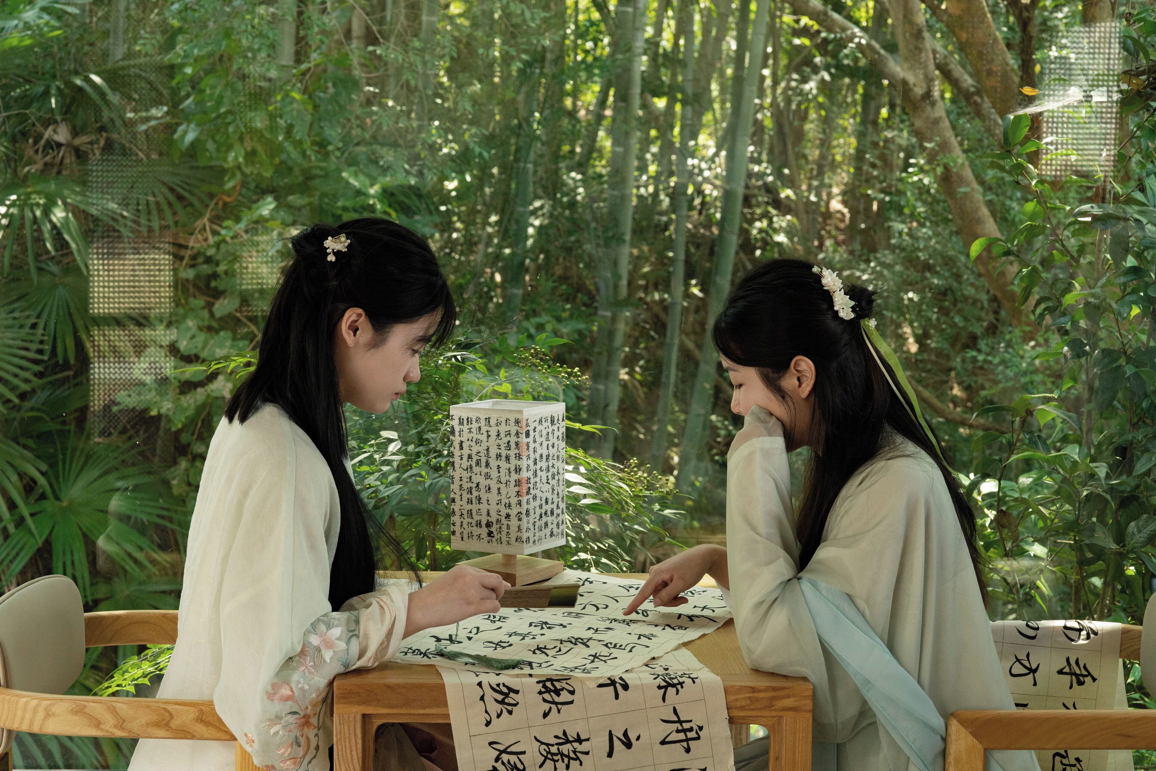 Two individuals seated at a table with a traditional Chinese calligraphy set, surrounded by lush greenery.