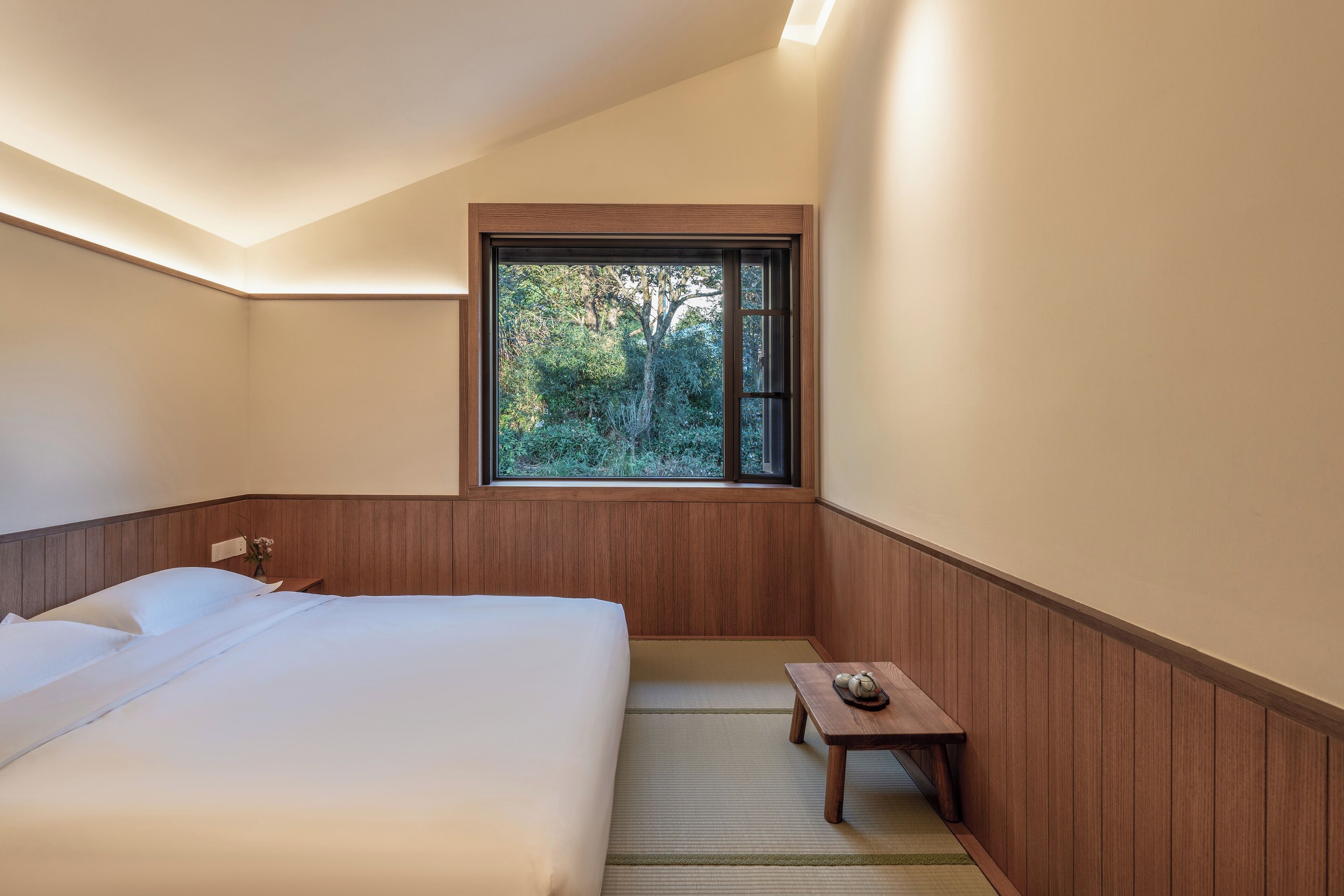A bedroom with a large bed, a small table with a tea set, a window with a view of trees, and wooden paneling on the walls.