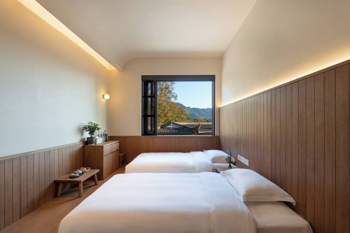 A modern hotel room with two beds, a small wooden bench, a nightstand, and a window with a view of mountains.