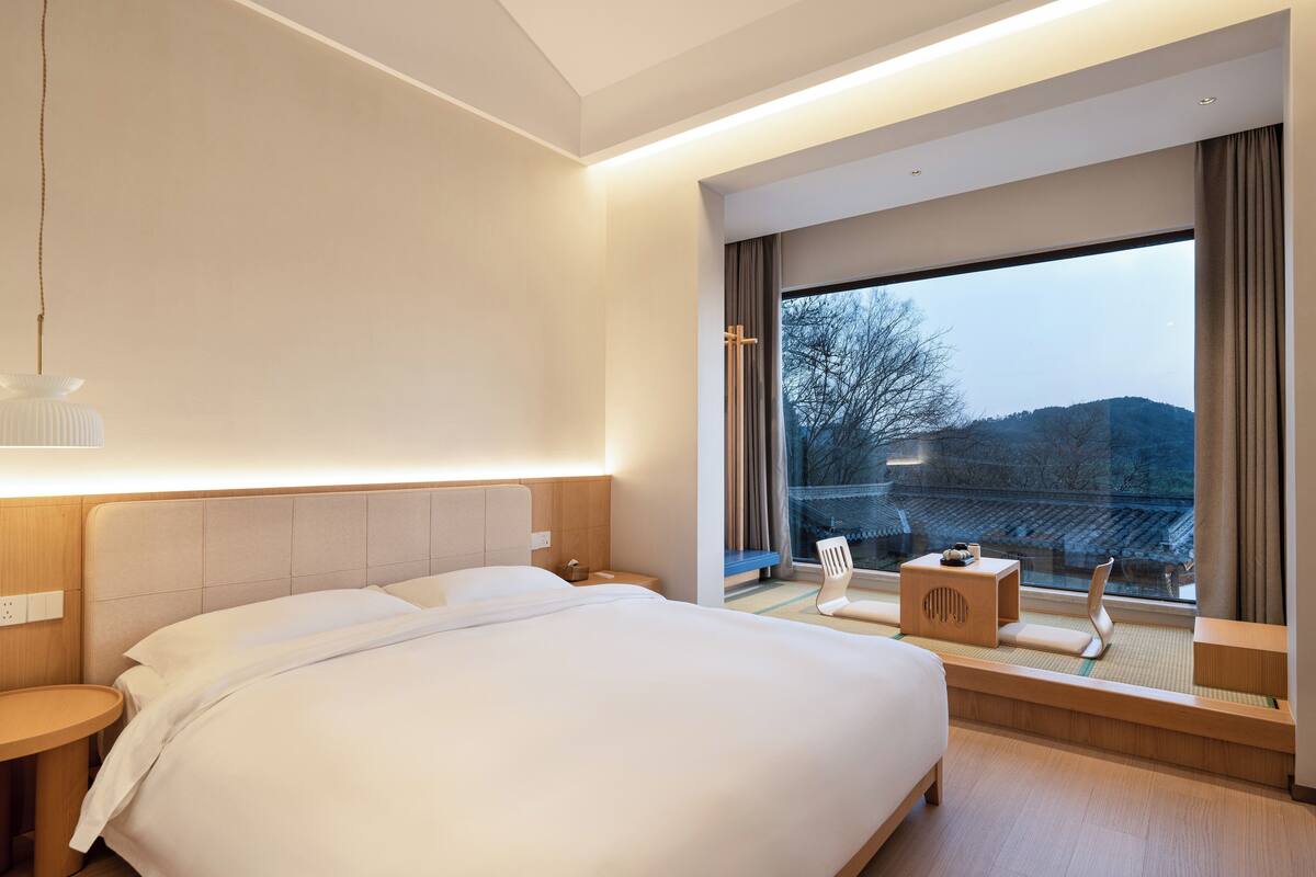 A modern hotel room with a large bed, a small side table, a chair, and a large window offering a view of the outdoors.