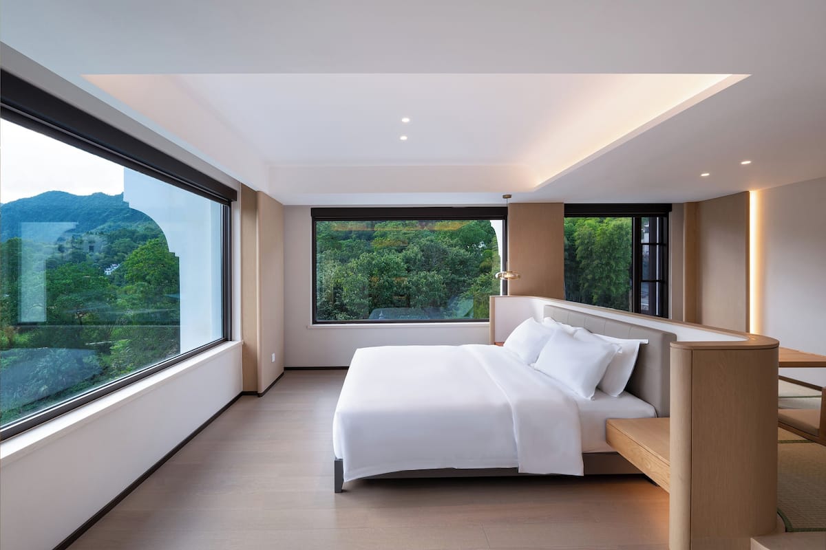 A modern bedroom with a large bed, a view of greenery, and a cityscape through large windows.