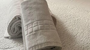 Una cama bien tendida con un edredón blanco texturizado, una toalla enrollada y un almohadón con diseño circular.