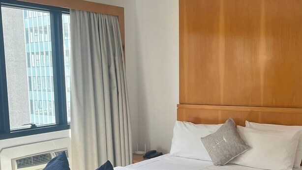 Habitación de hotel con cama, sofá, cabecero de madera, ventana con cortinas y vistas a edificios.