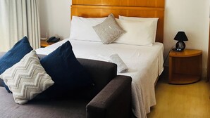Una habitación de hotel con cabecera de madera, cama con ropa de cama blanca, sofá gris y una mesita de noche con lámpara.