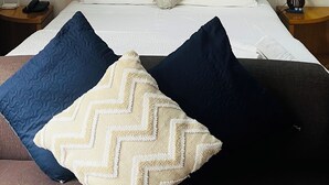 Una habitación de hotel con cabecera de madera, cama con almohadas blancas y oscuras, lámpara de noche y teléfono en la mesita de noche.