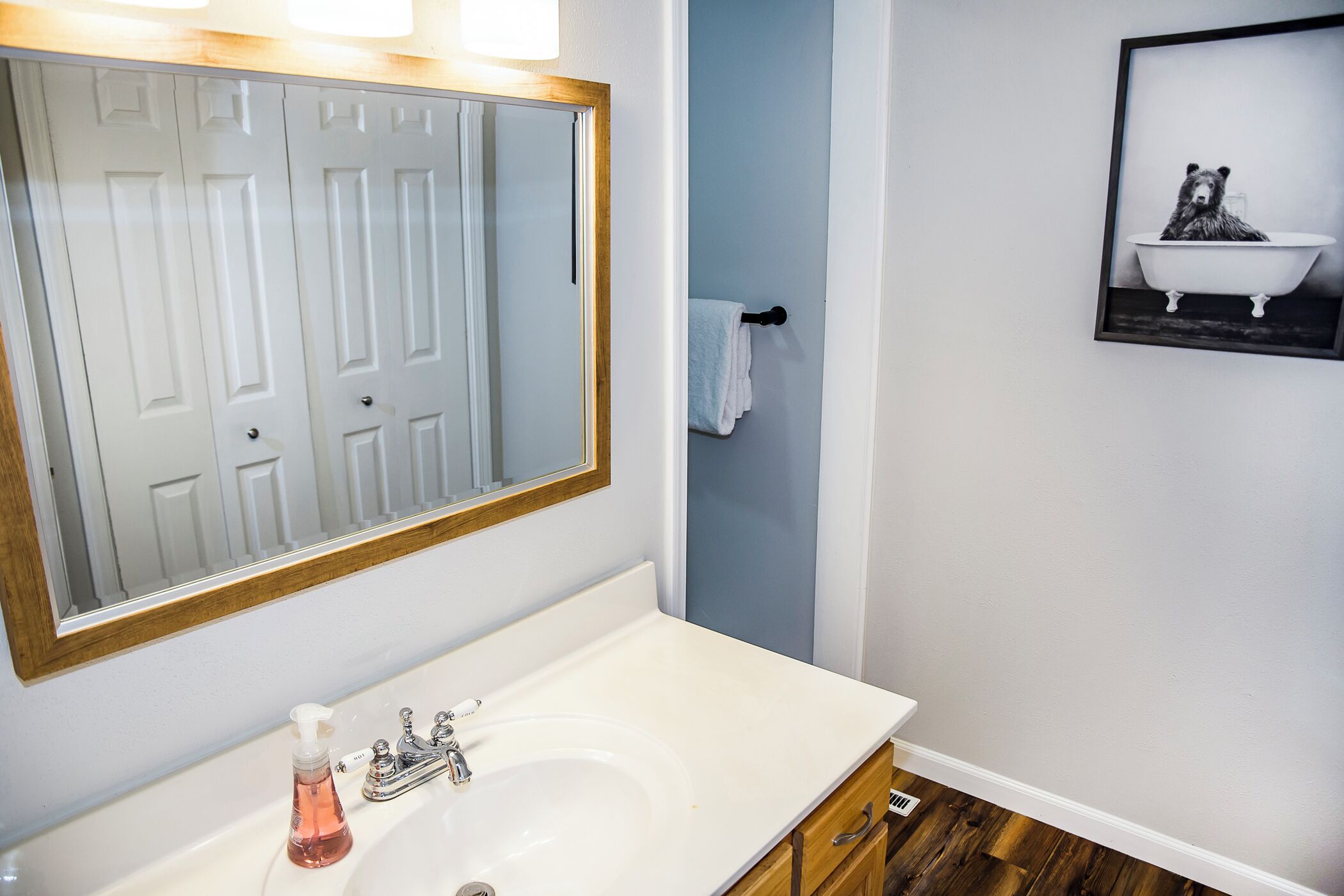 Une salle de bain avec un lavabo blanc, un miroir avec un cadre en bois, un tableau représentant un ours dans une baignoire, et une serviette suspendue à un porte-serviettes.
