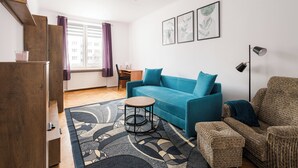 Una sala de estar moderna con un sofá azul, una mesita redonda pequeña, un tapete estampado y vistas de la ciudad a través de una ventana con cortinas moradas.
