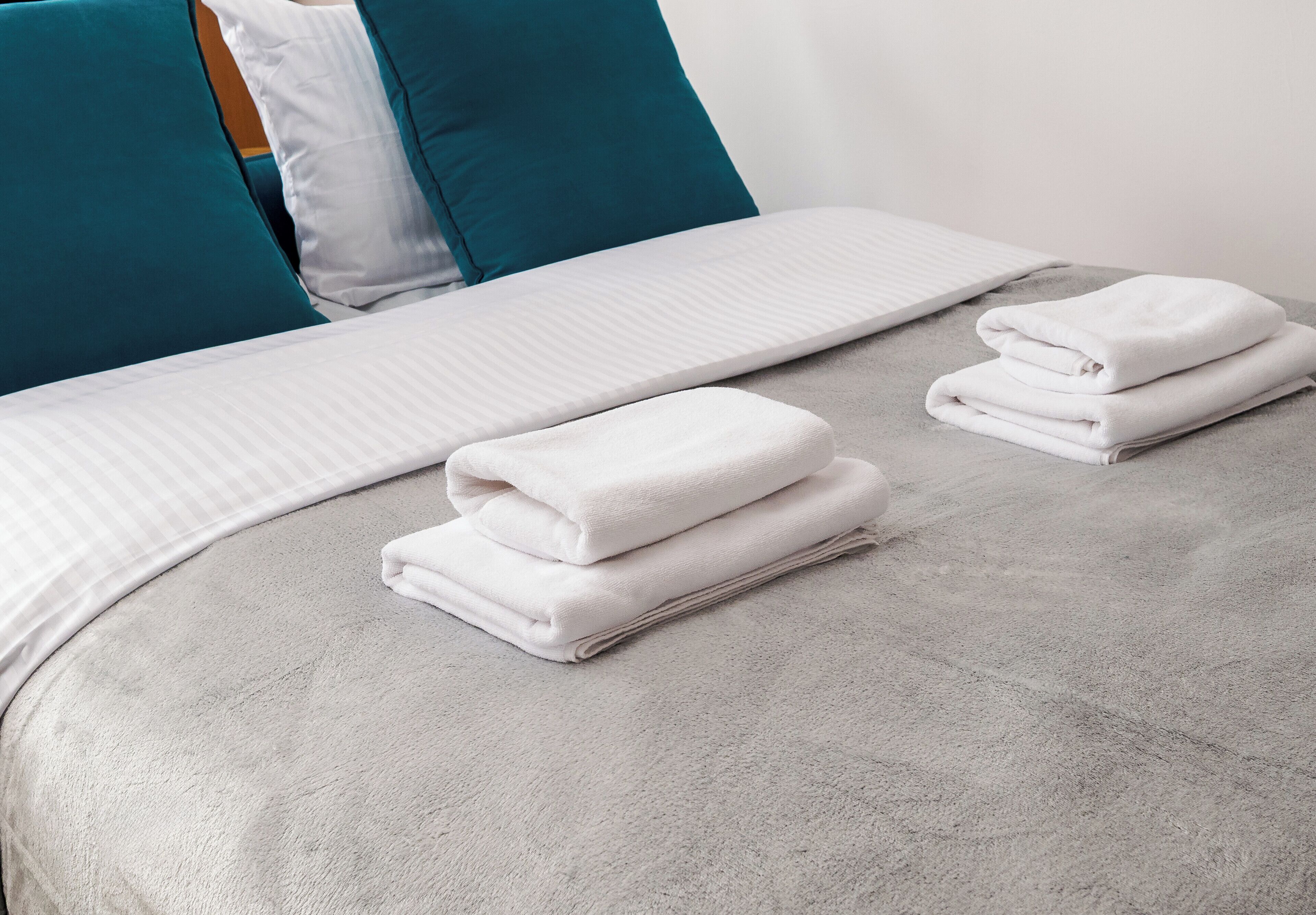 A neatly made bed with white linens and teal pillows, a folded white towel on the bed, and a grey carpeted floor.