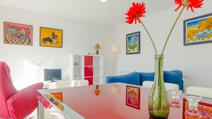 Ein modernes Wohnzimmer mit einem roten Sofa, einem Glastisch und einer grünen Vase mit roten Blumen. An den Wänden befinden sich Gemälde und im Hintergrund steht ein weißes Sideboard.