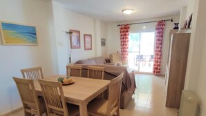 A dining area with a wooden table and chairs, a sofa, and a view of the outdoors through a large window with patterned curtains. - Rotonda VII 54 in Conil de la Frontera (Conil de la Frontera)