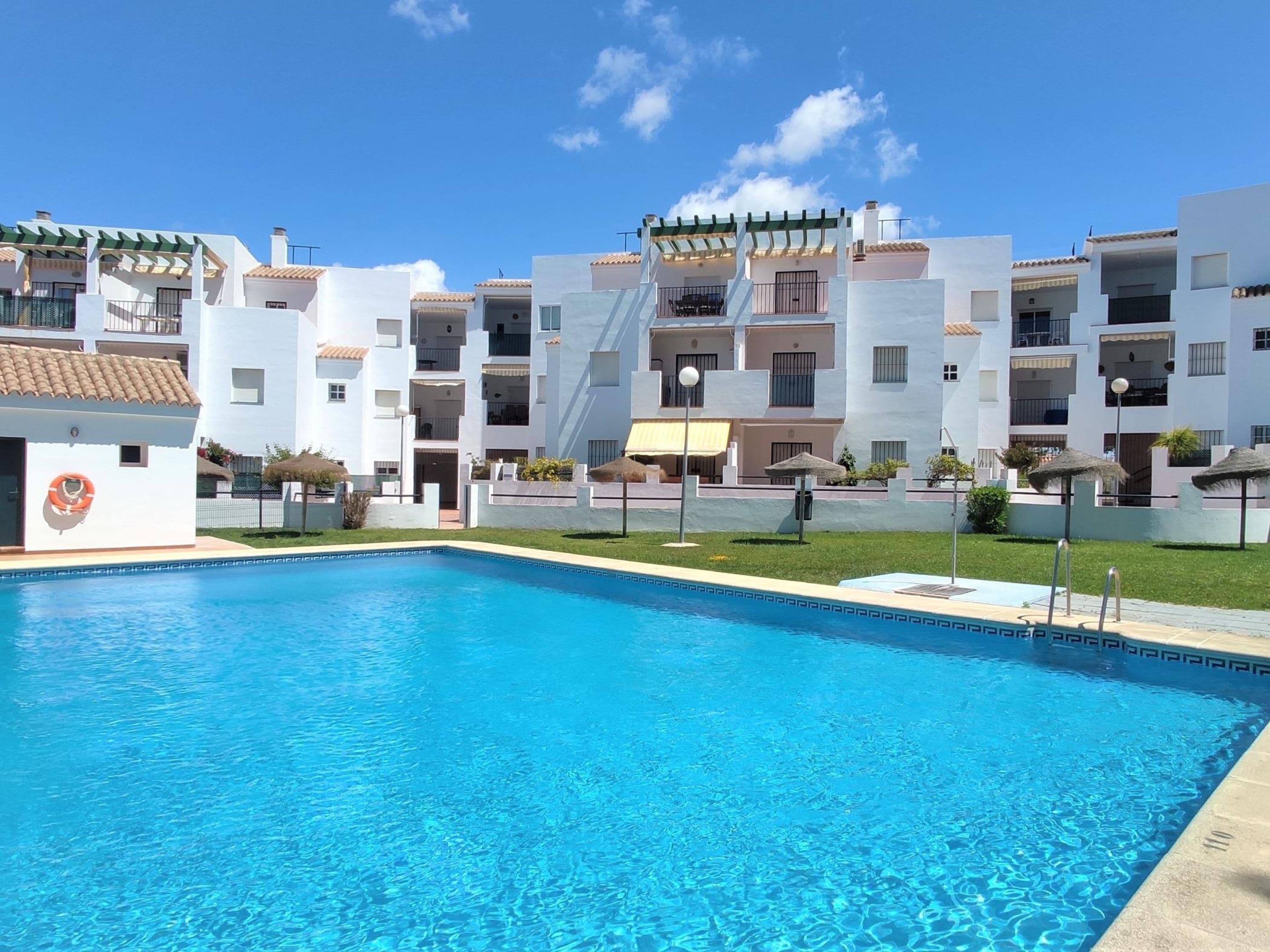 A swimming pool with a clear blue water surface, surrounded by white buildings with balconies and a covered outdoor seating area.