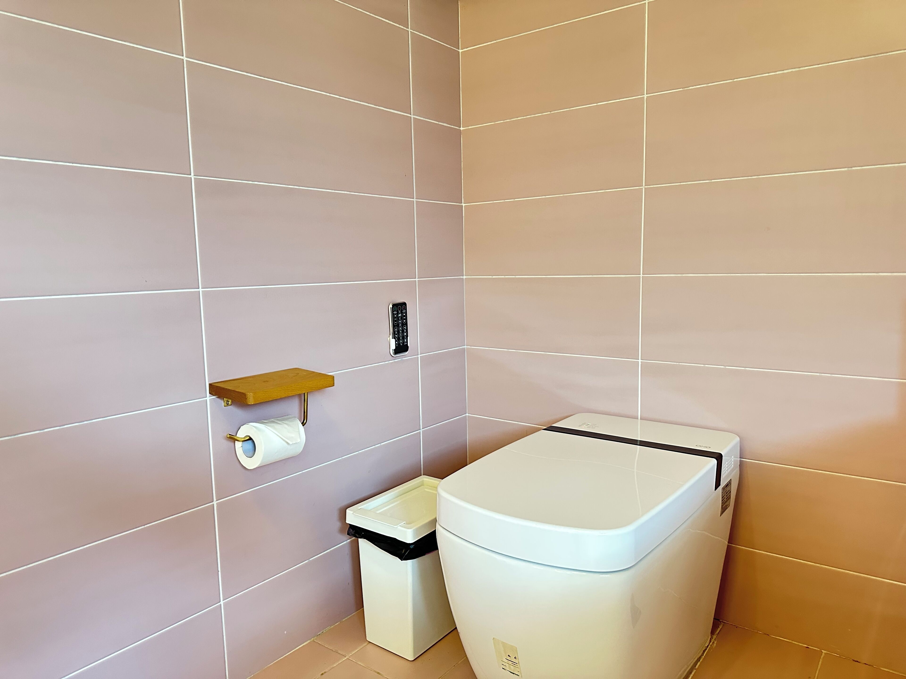 A bathroom with a toilet, a trash bin, a toilet paper holder, and a shelf.