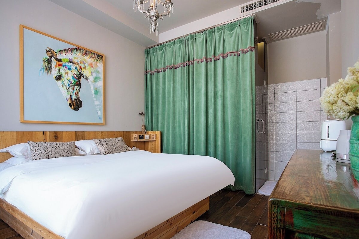 A bedroom with a wooden bed, a green curtain, a painting of a horse, and a bathroom with a white sink and a vase of flowers.