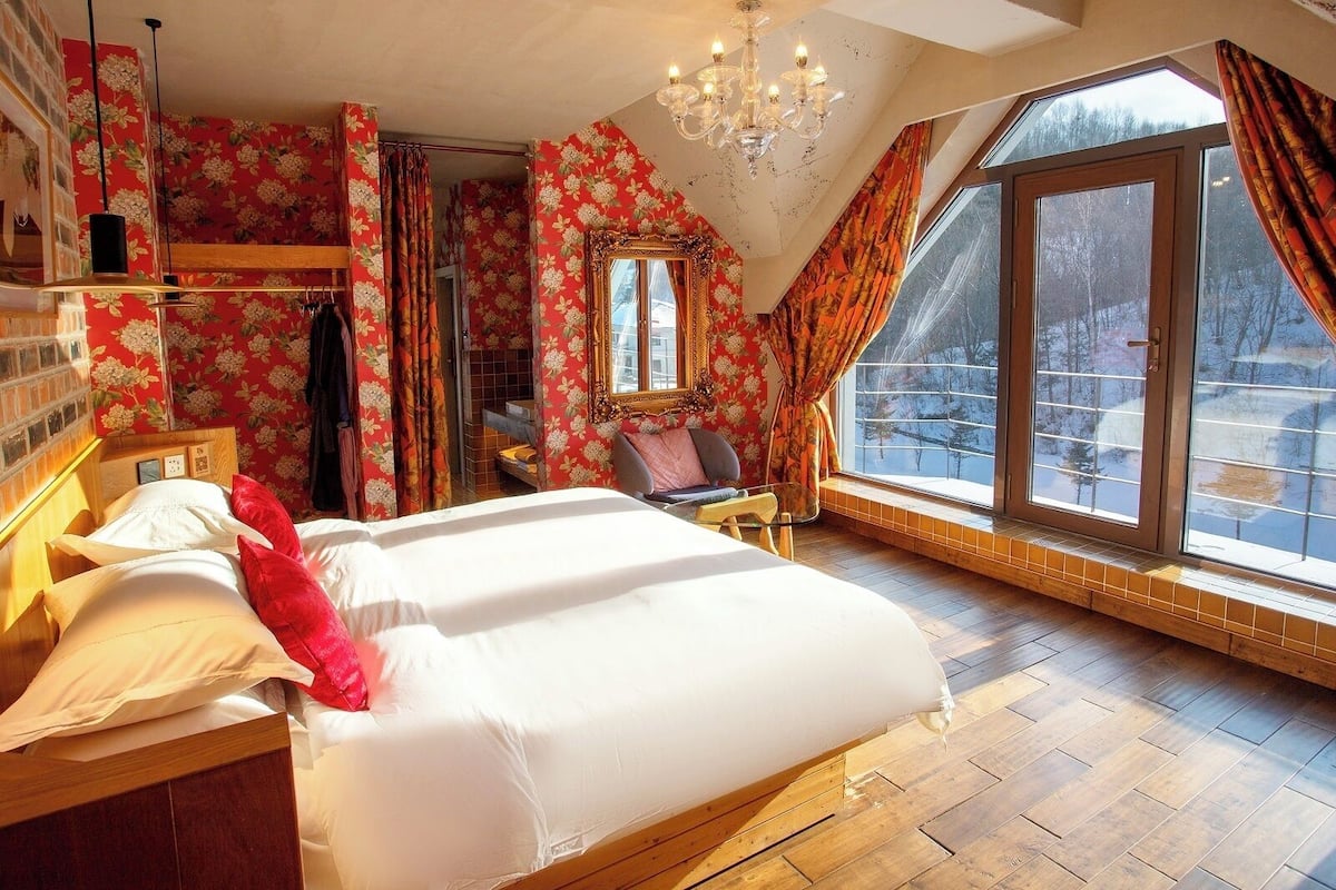 A bedroom with a large bed, a chandelier, a window with curtains, and a view of the outside.
