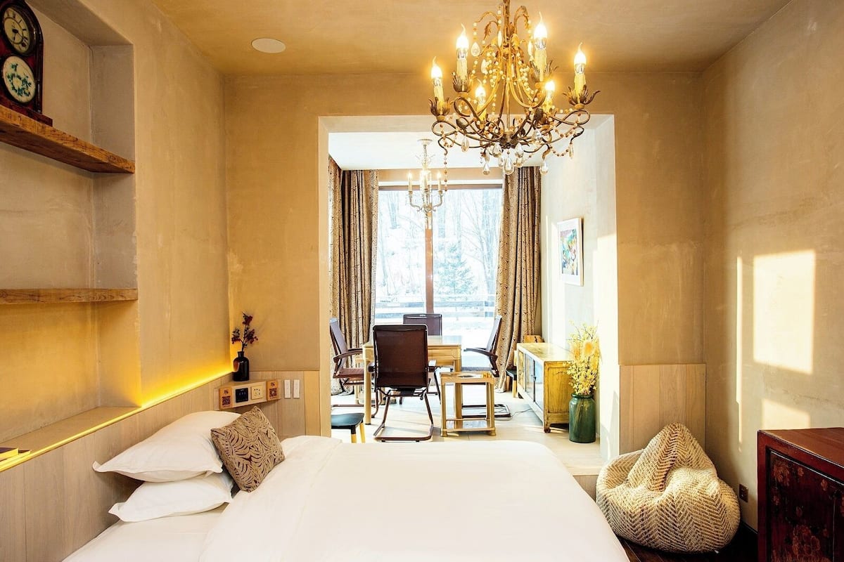 A hotel room with a large bed, a chandelier, a desk with chairs, and a view of the outdoors.